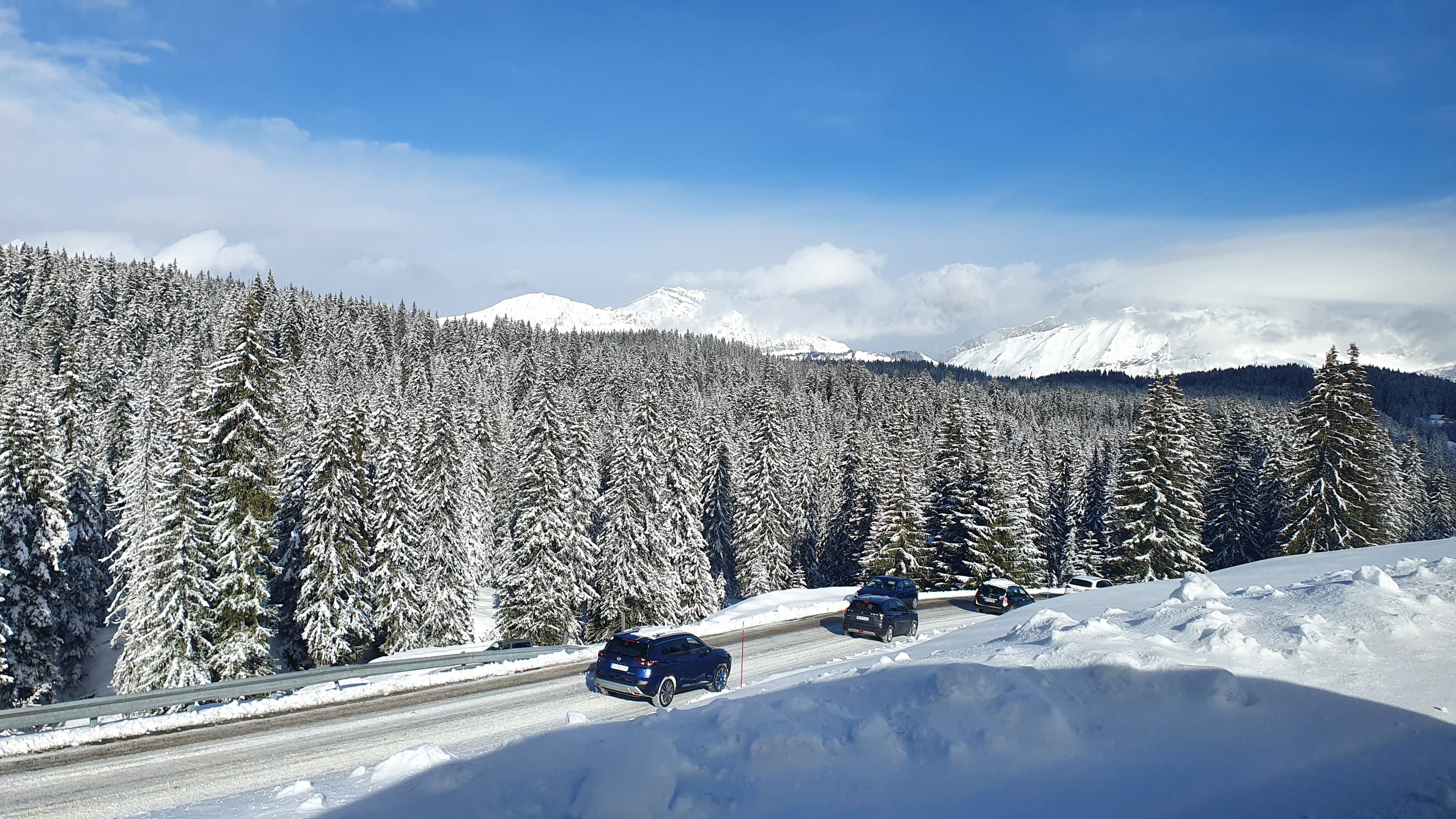 Vue d'une route de montagne enneigée sous le soleil avec quelques voitures circulant normalement lors du premier week-end des vacances d'hiver 2026.