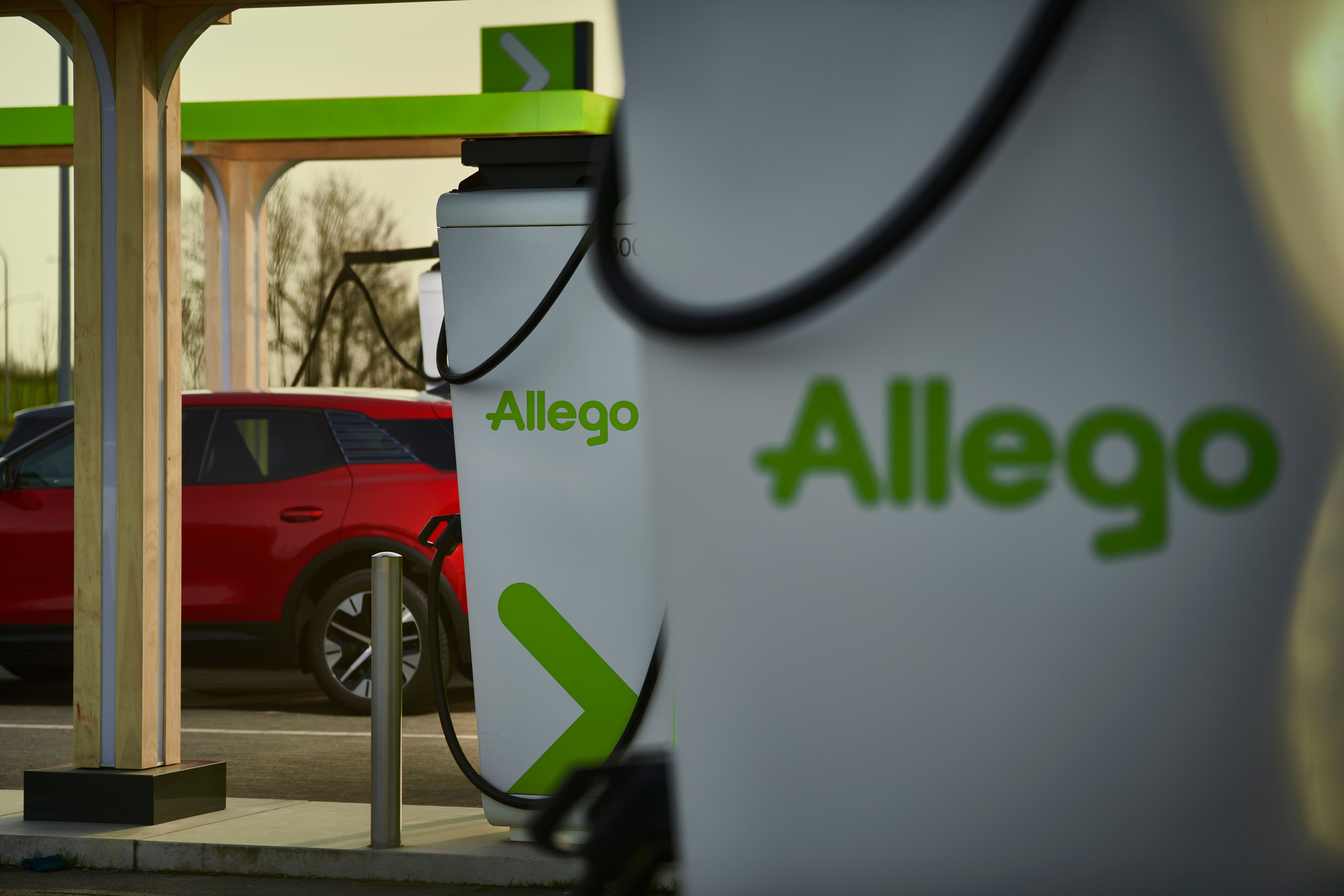 Gros plan sur une borne de recharge électrique Allego avec une voiture électrique rouge en arrière-plan
