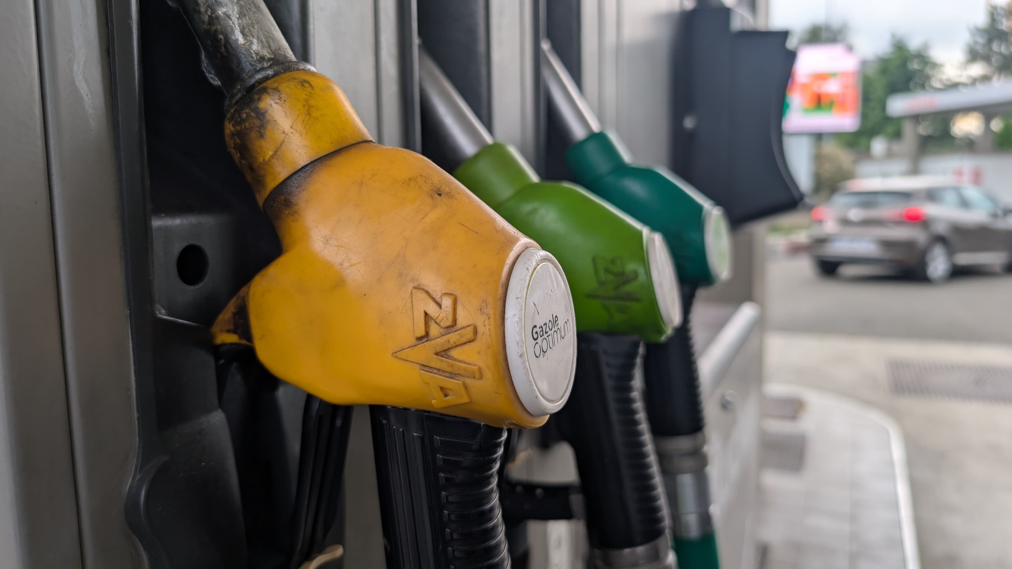 Zoom sur des pistolets de carburant dans une station service, une voiture circule en arrière-plan