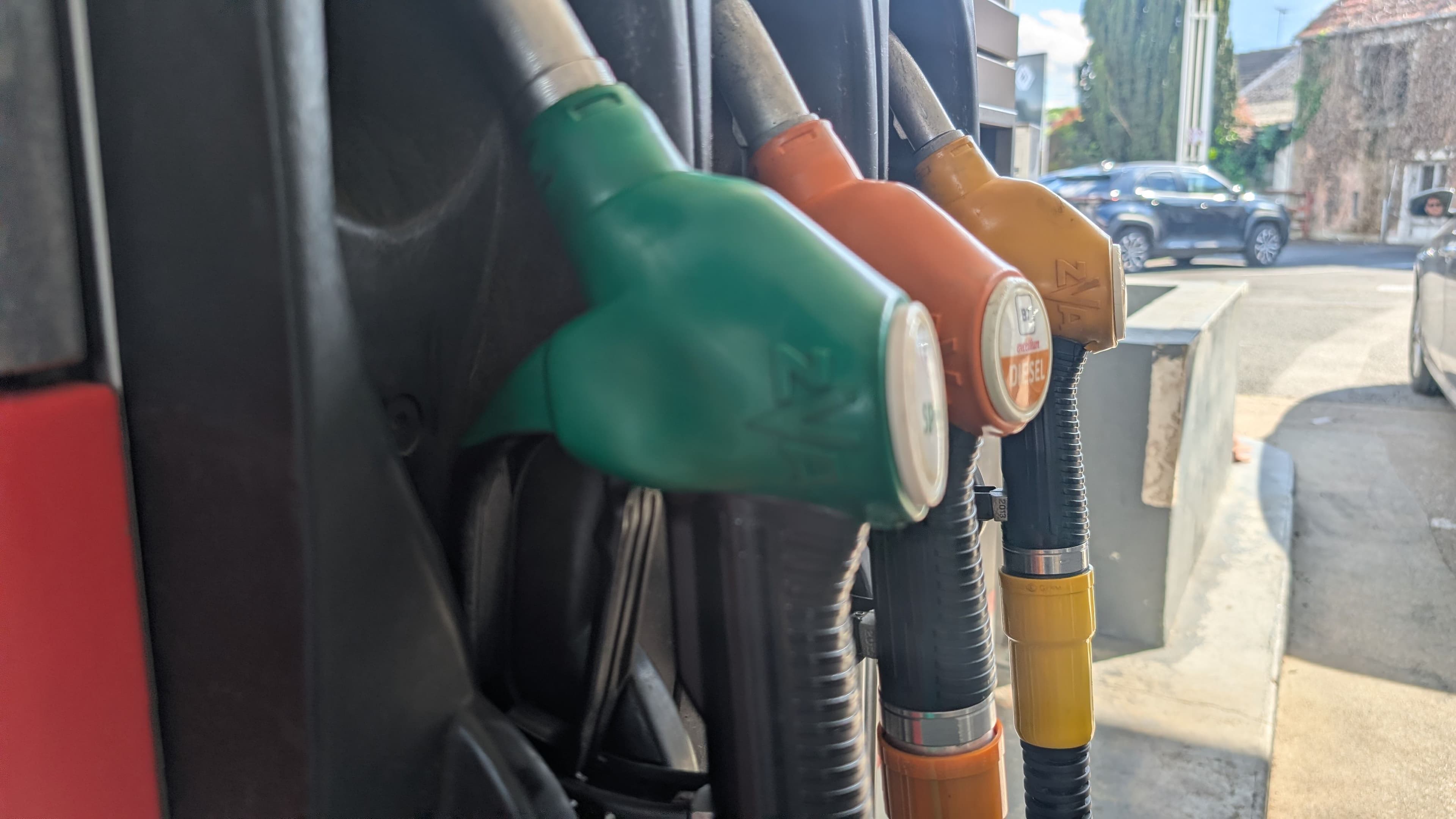 Une station essence avec les pistolets des différents carburants disponible (SP95 et gasoil) et une voiture quittant la station service en arrière plan