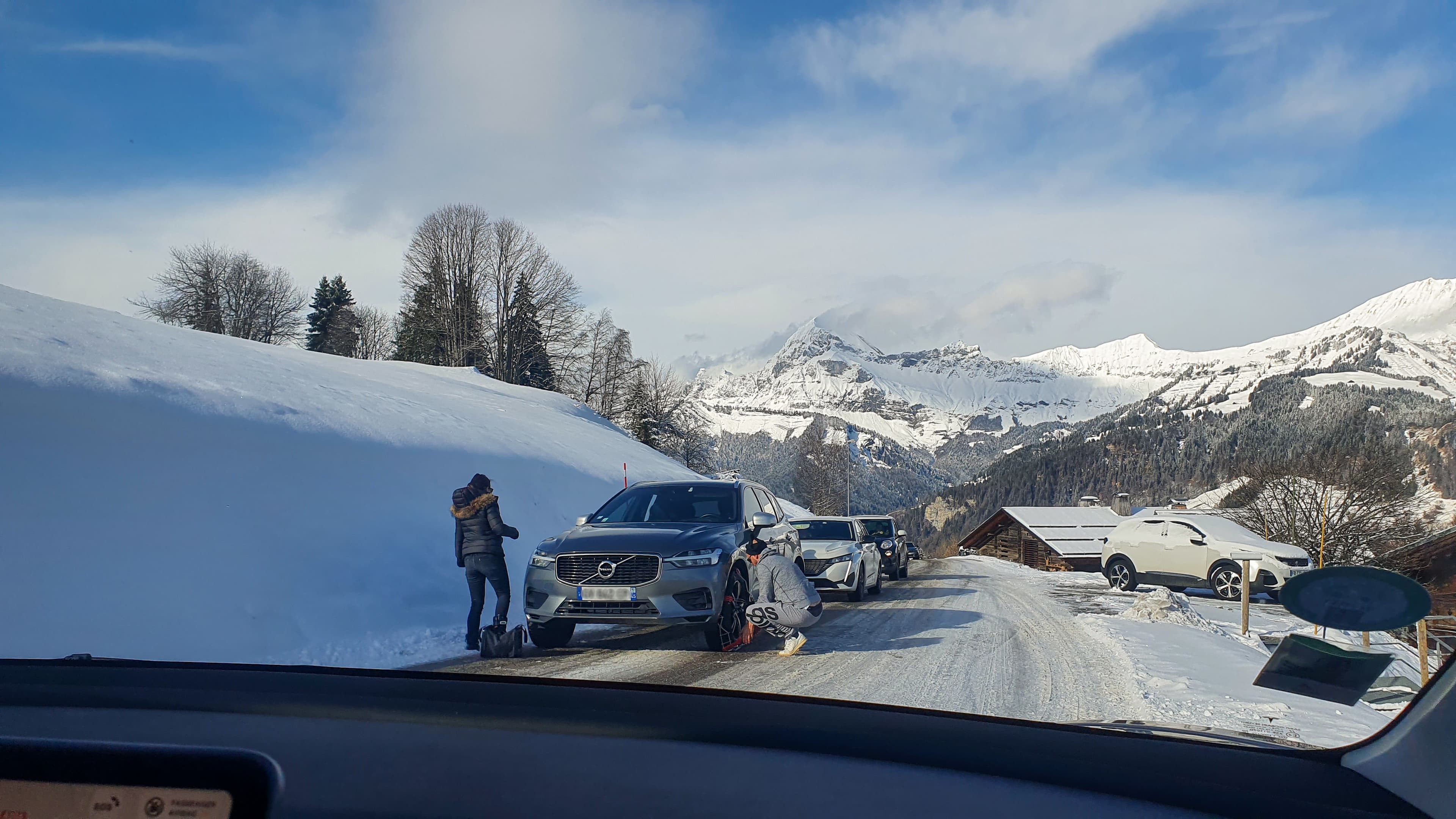 Automobiliste accroupi en train d’enfiler une chaussette à neige sur la roue avant de sa voiture, sur une route de montagne enneigée
