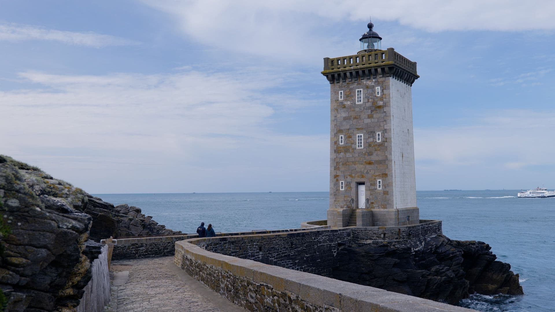 Phare en pierre au bord de l’océan avec passerelle et bateaux au loin