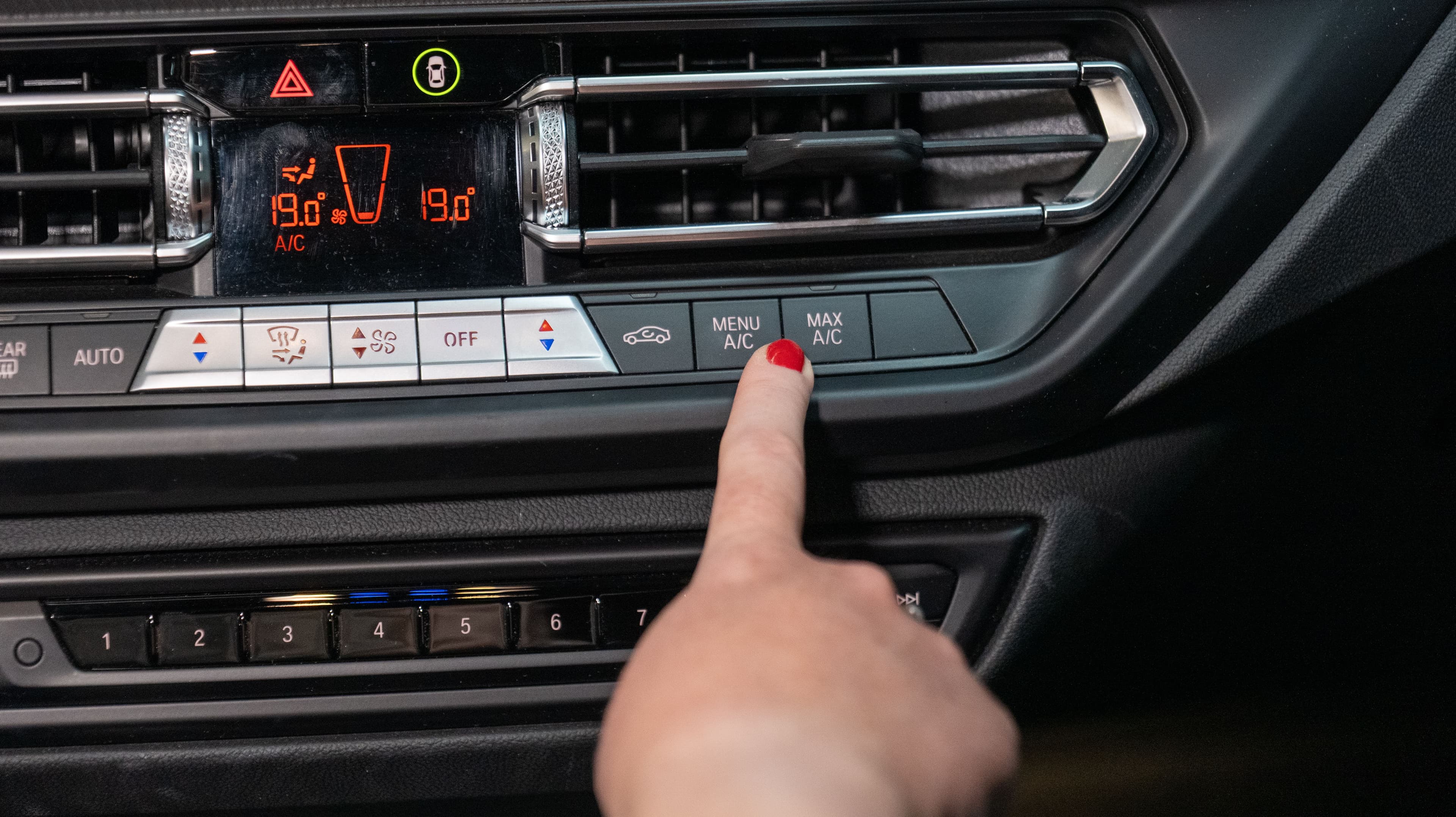 Une main posée sur le bouton "Max AC" du tableau de bord pour régler la climatisation. 