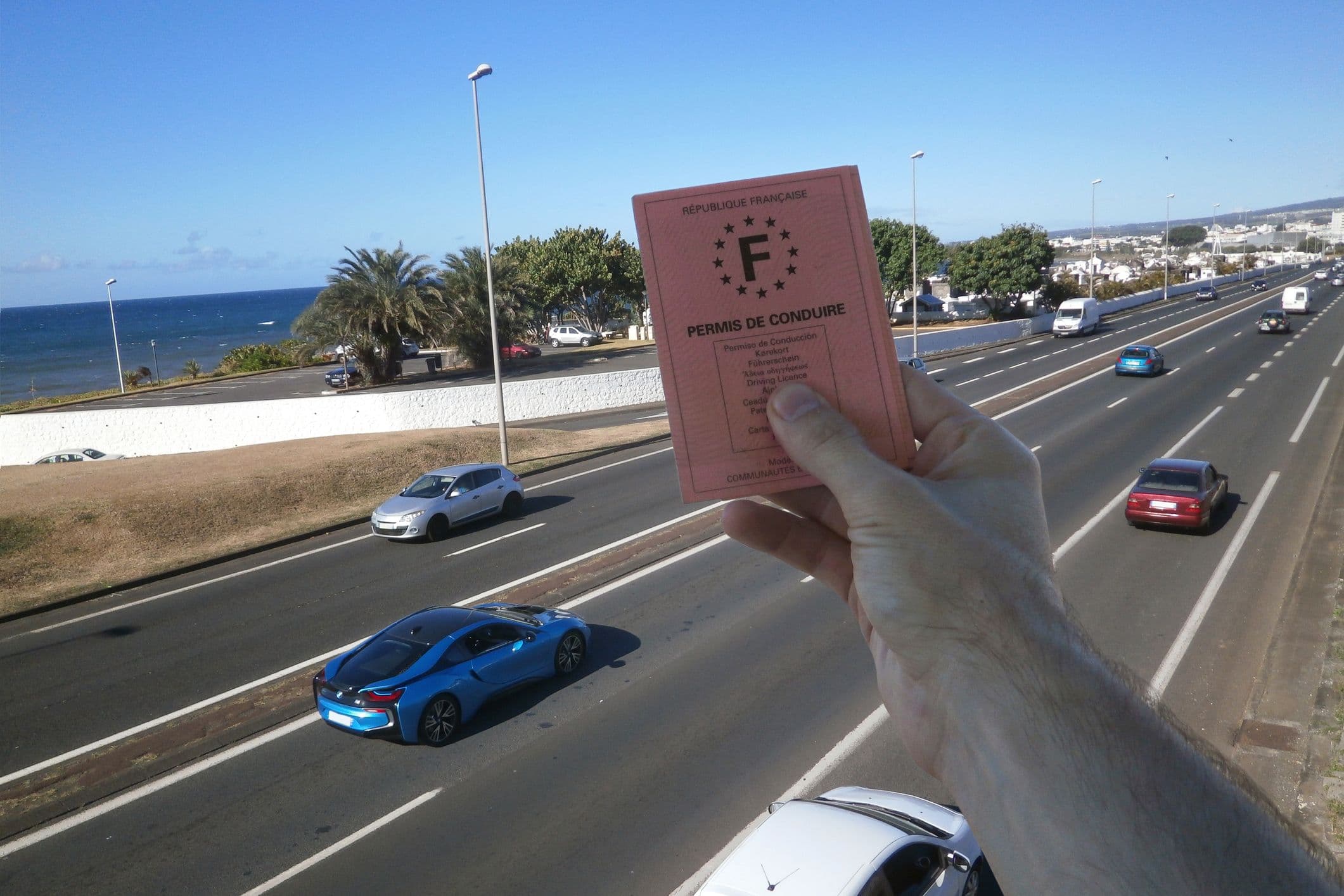 Permis de conduire français tenu au-dessus d’une route côtière avec voitures étrangères