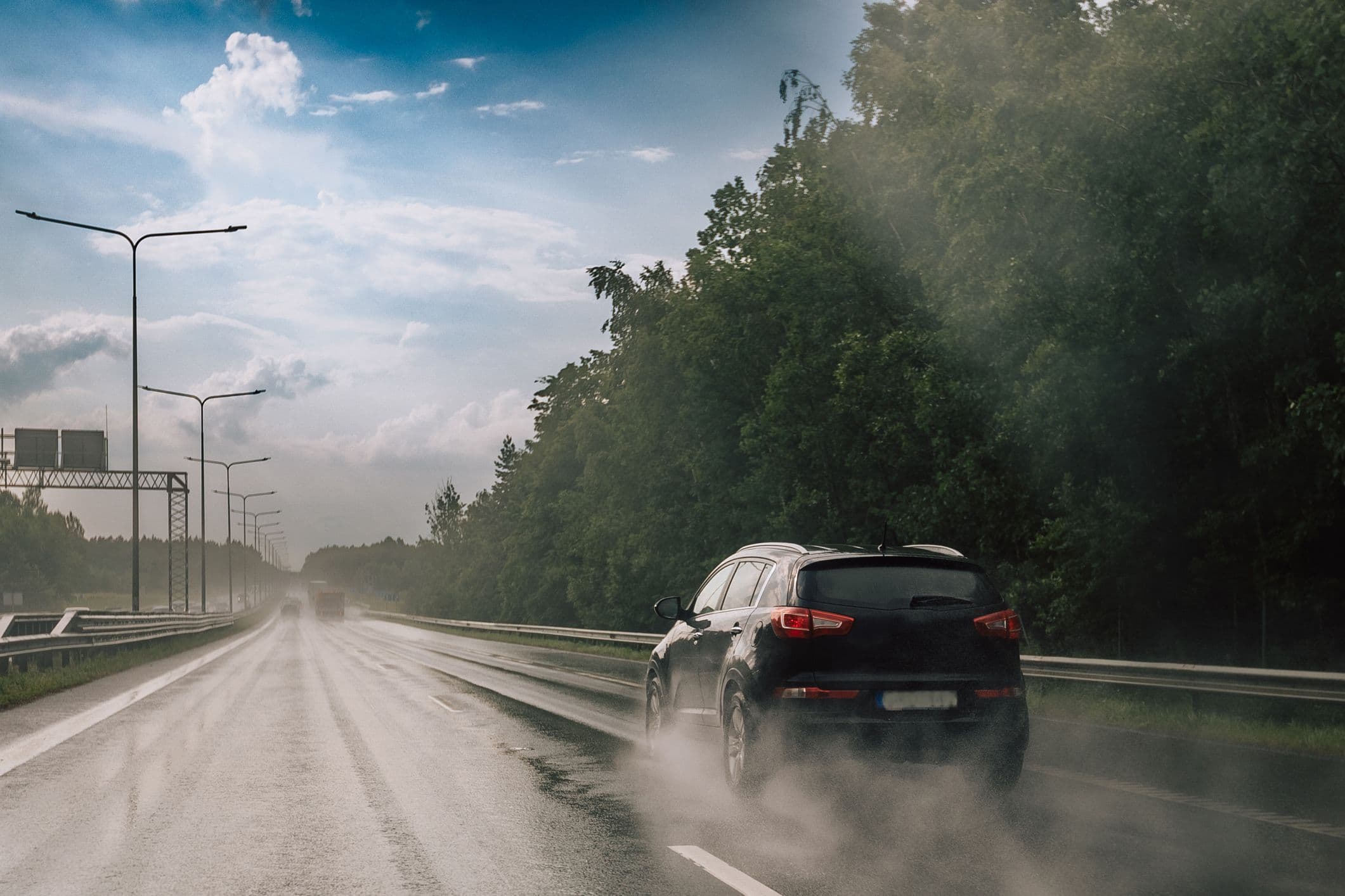 Une voiture noire circule sur une route mouillée sous un ciel bleu légèrement nuageux. La route est bordé d'arbres verts.