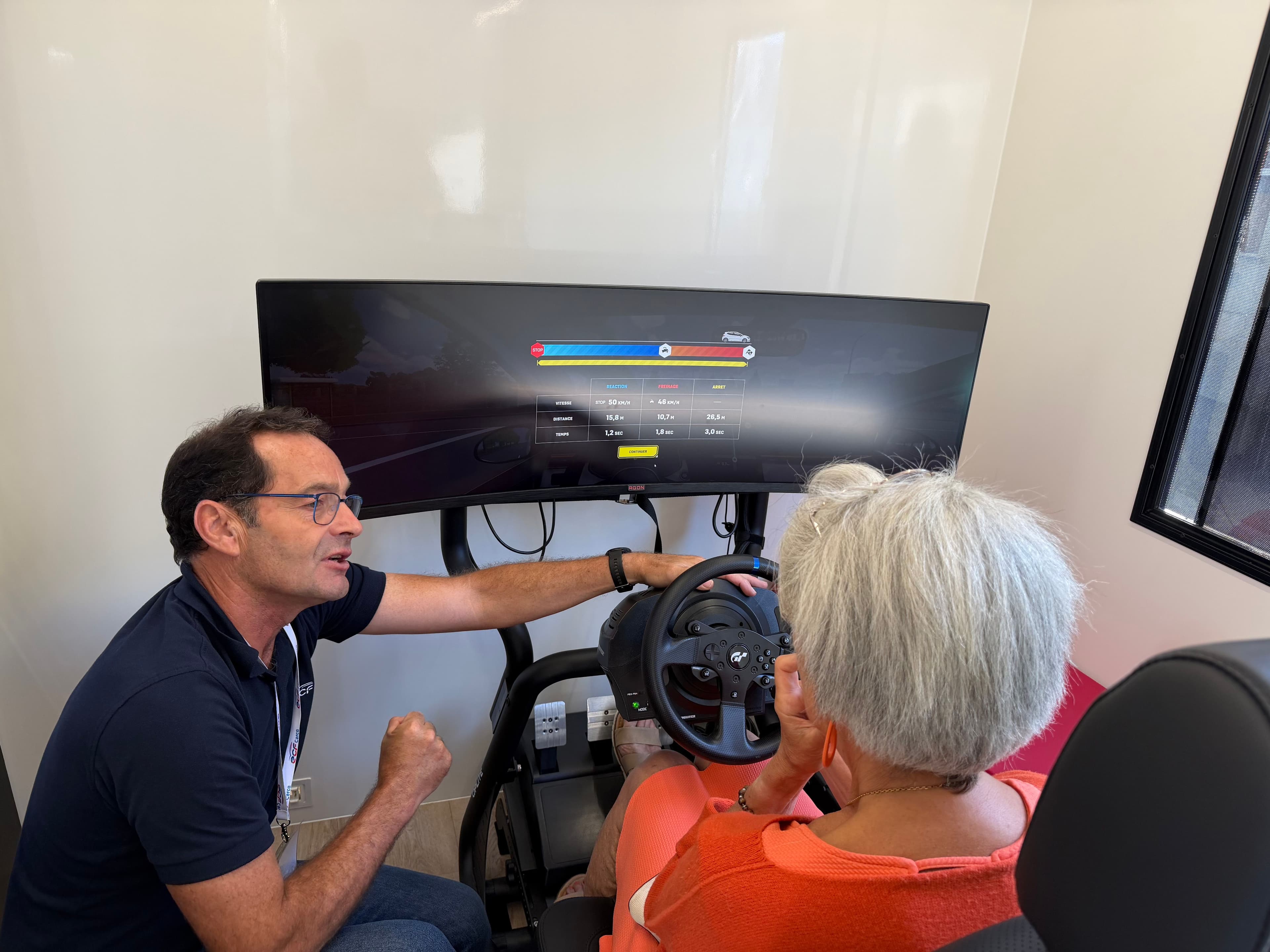 Une femme senior sur un simulateur de conduite, accompagnée d'un moniteur à la conduite. 
