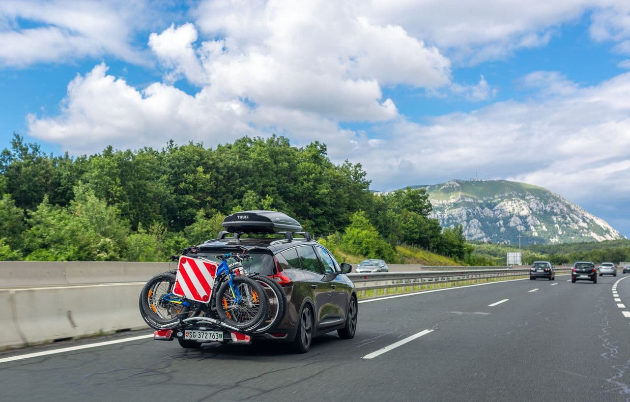 une voiture roule sur autoroute avec un coffre de toit et plusieurs vélo arrimés à l'arrière