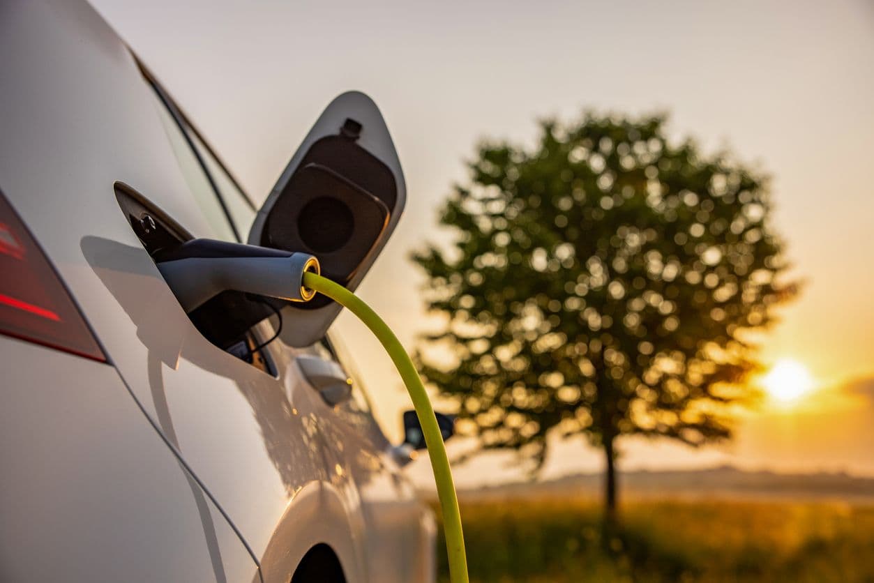 Une voiture électrique est en train de se recharger au coucher du soleil devant un arbre.