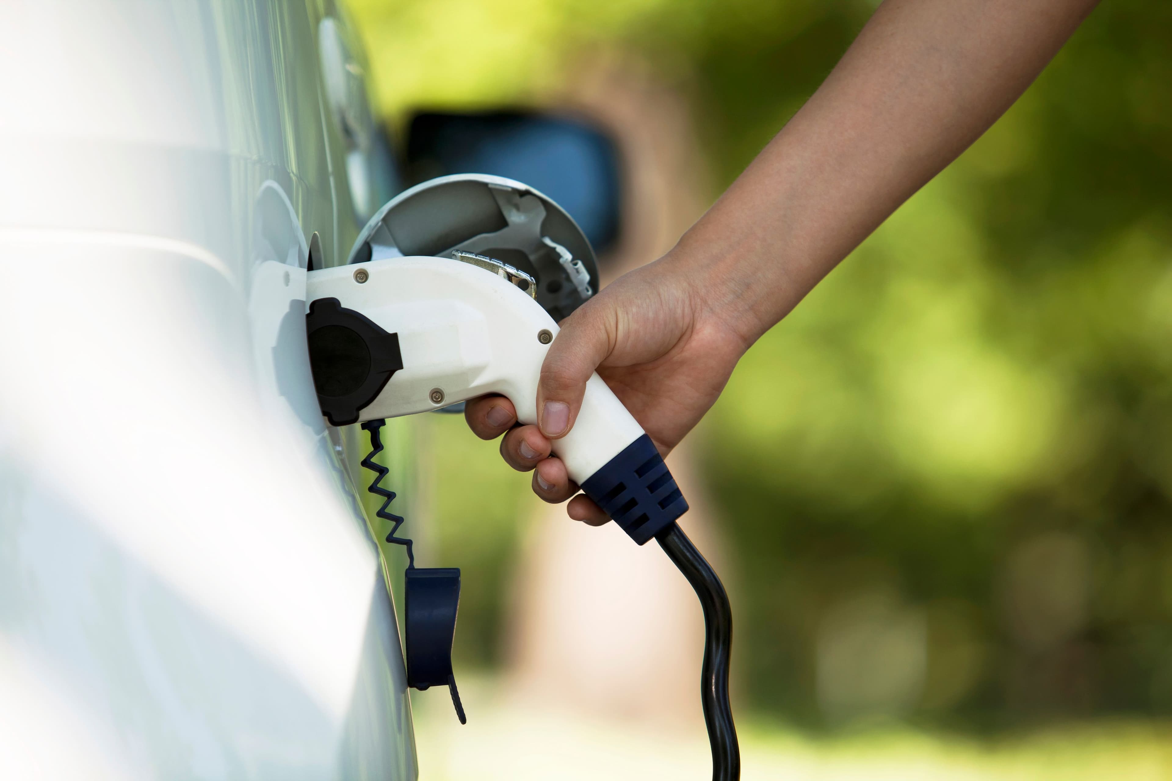 Une main tient un cable de recharge électrique vers la prise d'une voiture pour la recharger