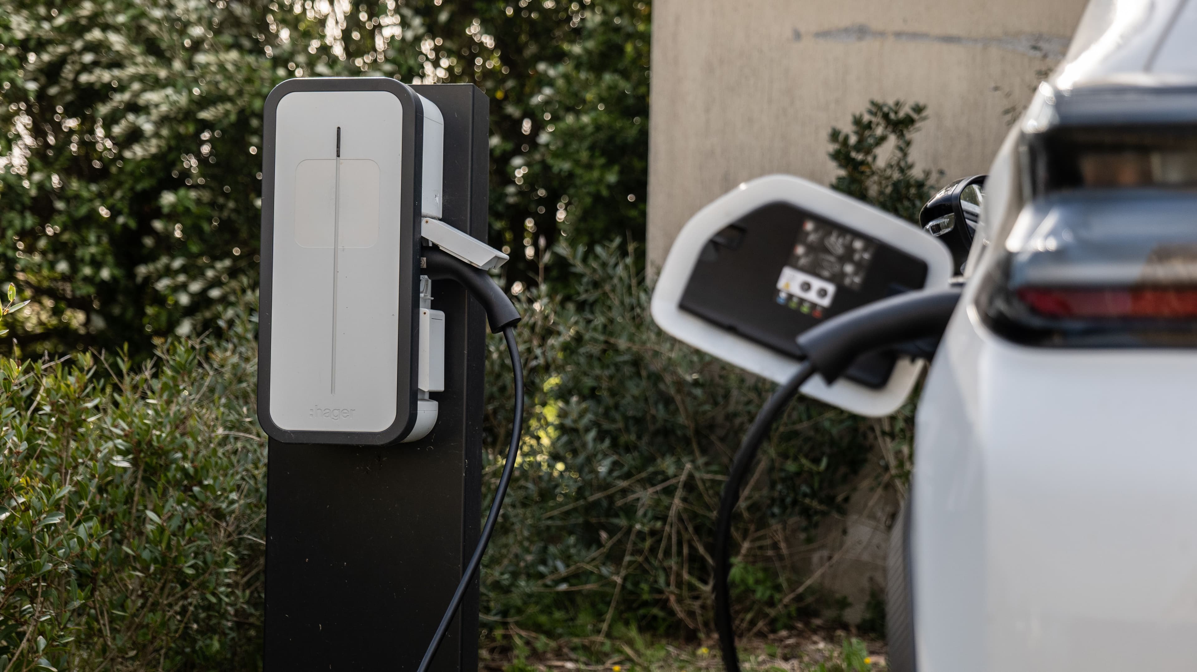 une voiture électrique blanche en train de se recharger sur une borne à domicile dans un jardinvégétalisé.