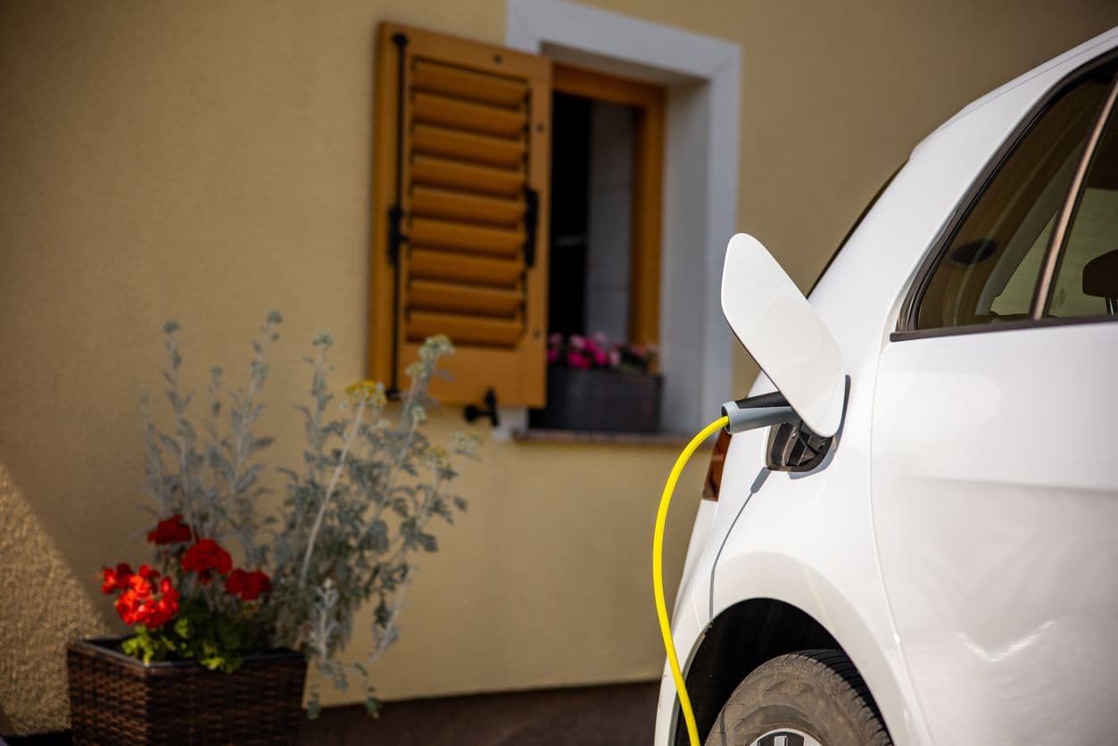Voiture électrique blanche en charge devant une maison avec fleurissement
