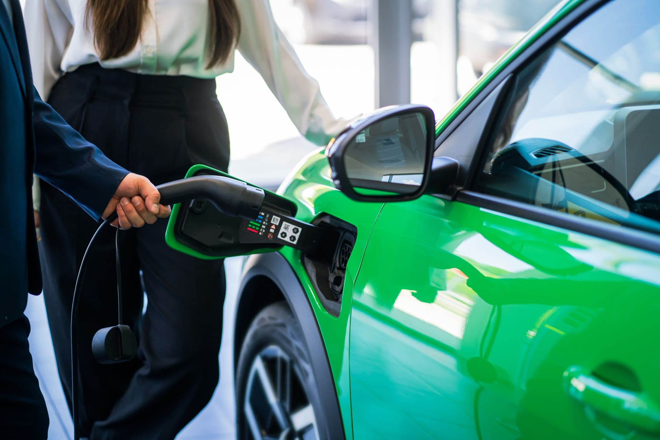 Un homme est en train de brancher une voiture électrique de couleur verte. Une femme appuie sa main sur le capot de la voiture.