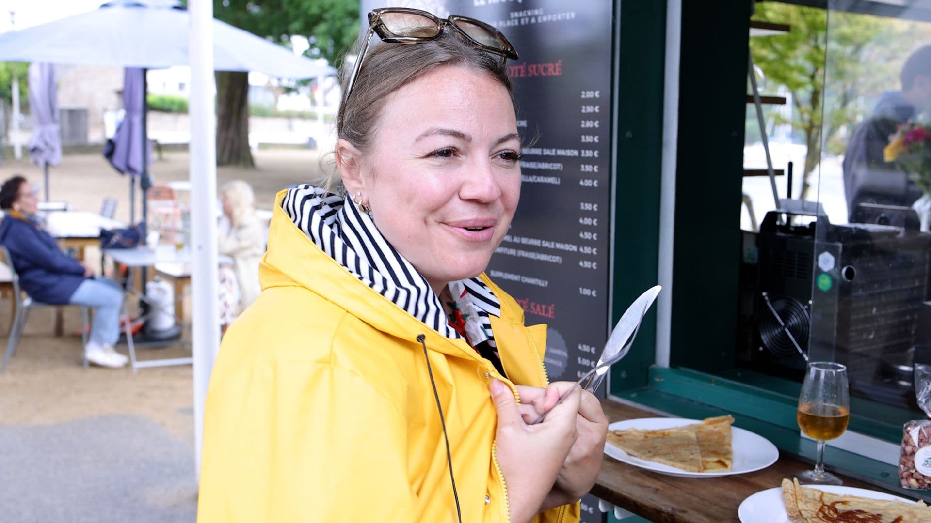 Pauline la journaliste mange des crêpes devant le comptoir du Kiosque de Mimi face à la caméra et vêtue d’une veste imperméable jaune typique de la Bretagne.